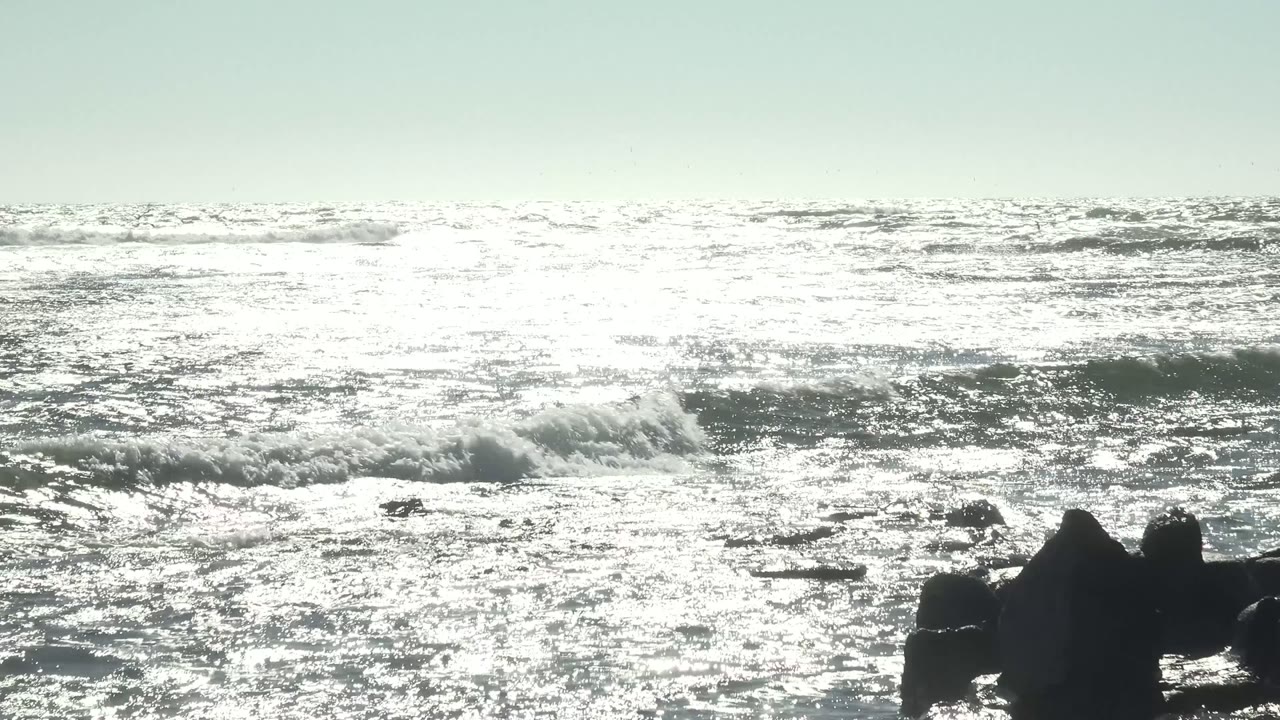 Sunlit water glitters across the frame at La Bocana, Baja California Sur, while whitecaps form behind low dark rocks and no visible people appear.