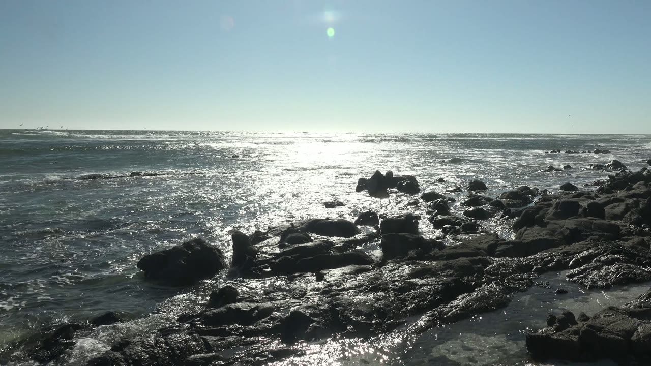 Wide view of dark rocks along the La Bocana shoreline with bright sun glare across the ocean and no visible people.