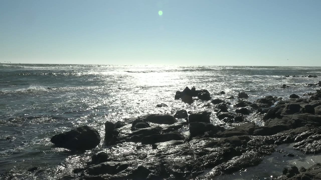 Ocean waves moving around dark rocks under a clear sky.