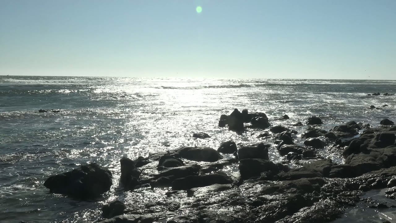 A low coastal composition at La Bocana shows silhouetted rocks in the foreground, rippled ocean in the middle distance, and a pale clear sky with no visible people.