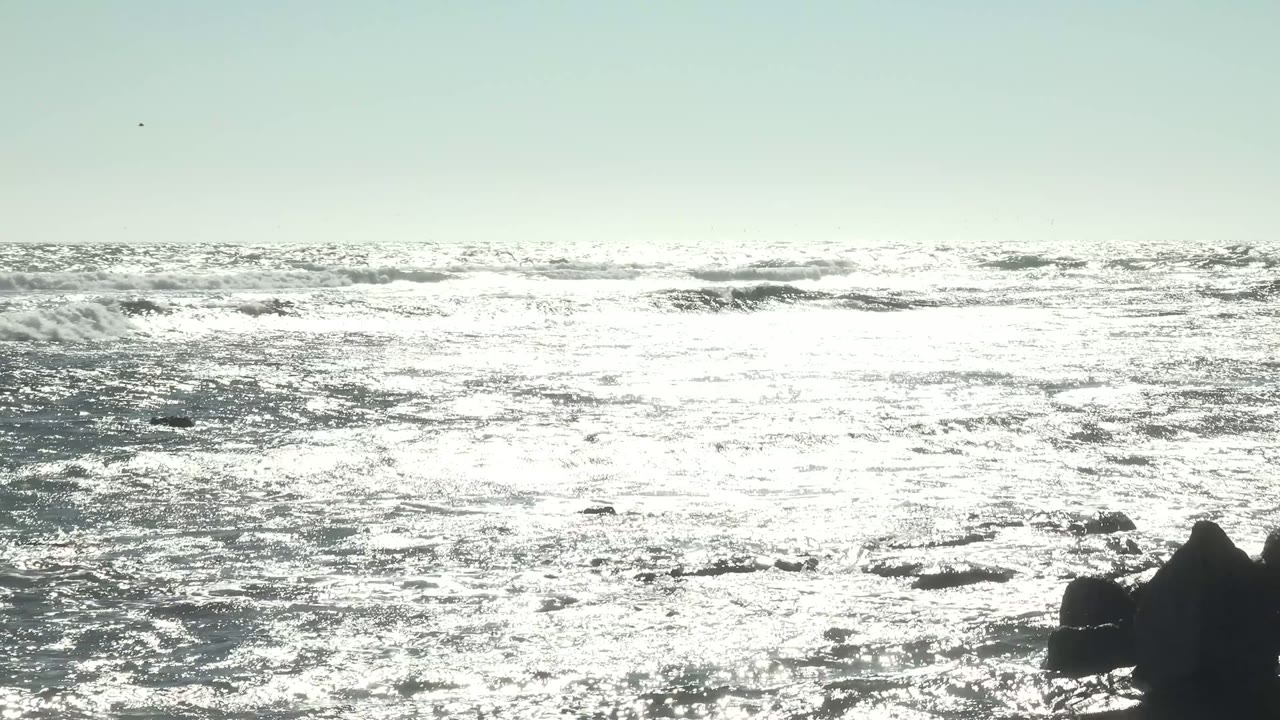 Sunlit ocean off La Bocana, Baja California Sur, with bright glare across the water, dark rocks near the shore, and no visible people.