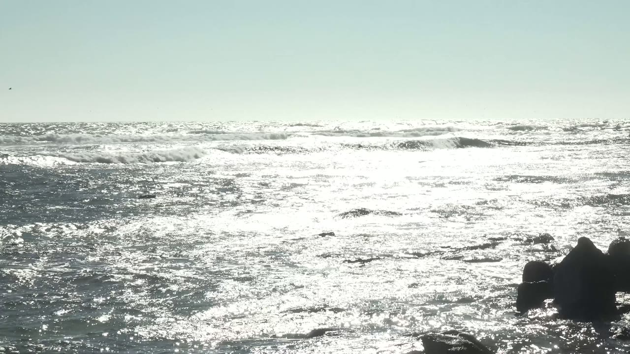 Bright ocean surface with waves breaking around shoreline rocks.