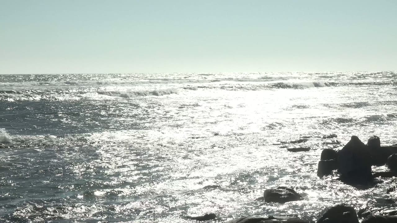 A wide La Bocana ocean view with intense sunlight on the water, low waves across the horizon, rocks at the right edge, and no visible people.