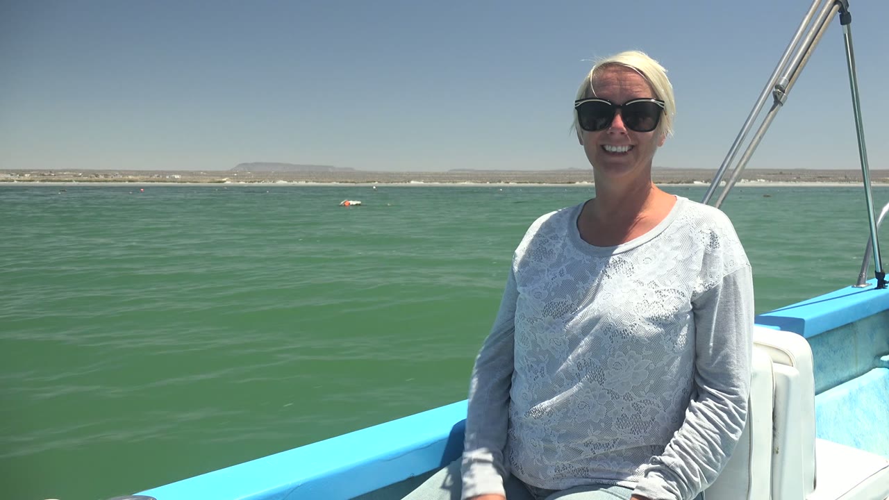 In La Bocana, Mexico, a woman in dark sunglasses and a gray long-sleeve shirt sits on a blue-and-white boat with green ocean water and a low shore behind her.