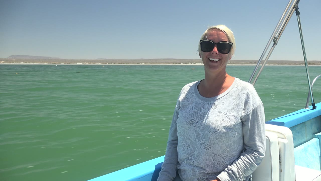 In La Bocana, Mexico, the woman sits near the right side of the frame on the blue-and-white boat as green water fills the middle ground and the low coast stretches behind her.