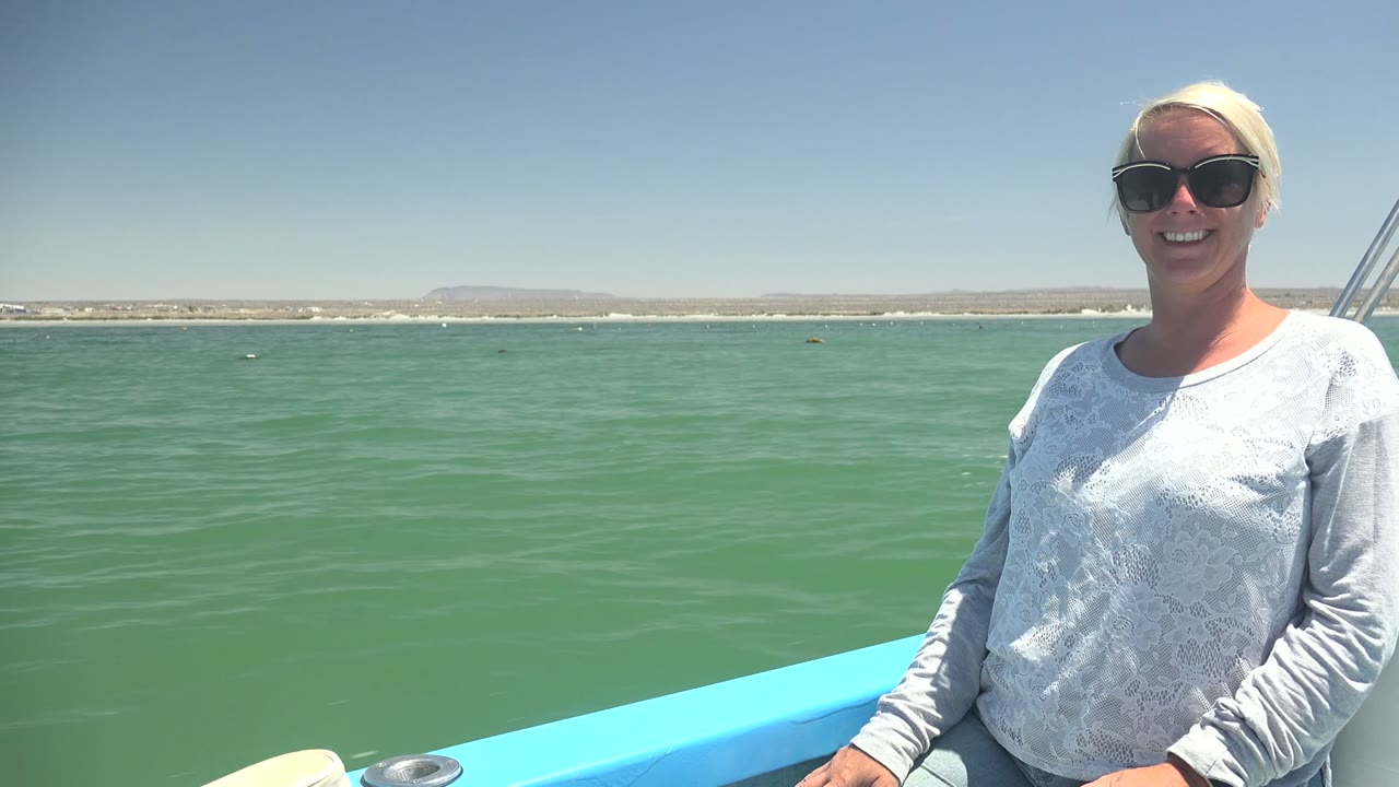 A blonde woman in sunglasses sits on a blue boat in La Bocana, Mexico, with calm green water, distant shore, and bright sky behind her.