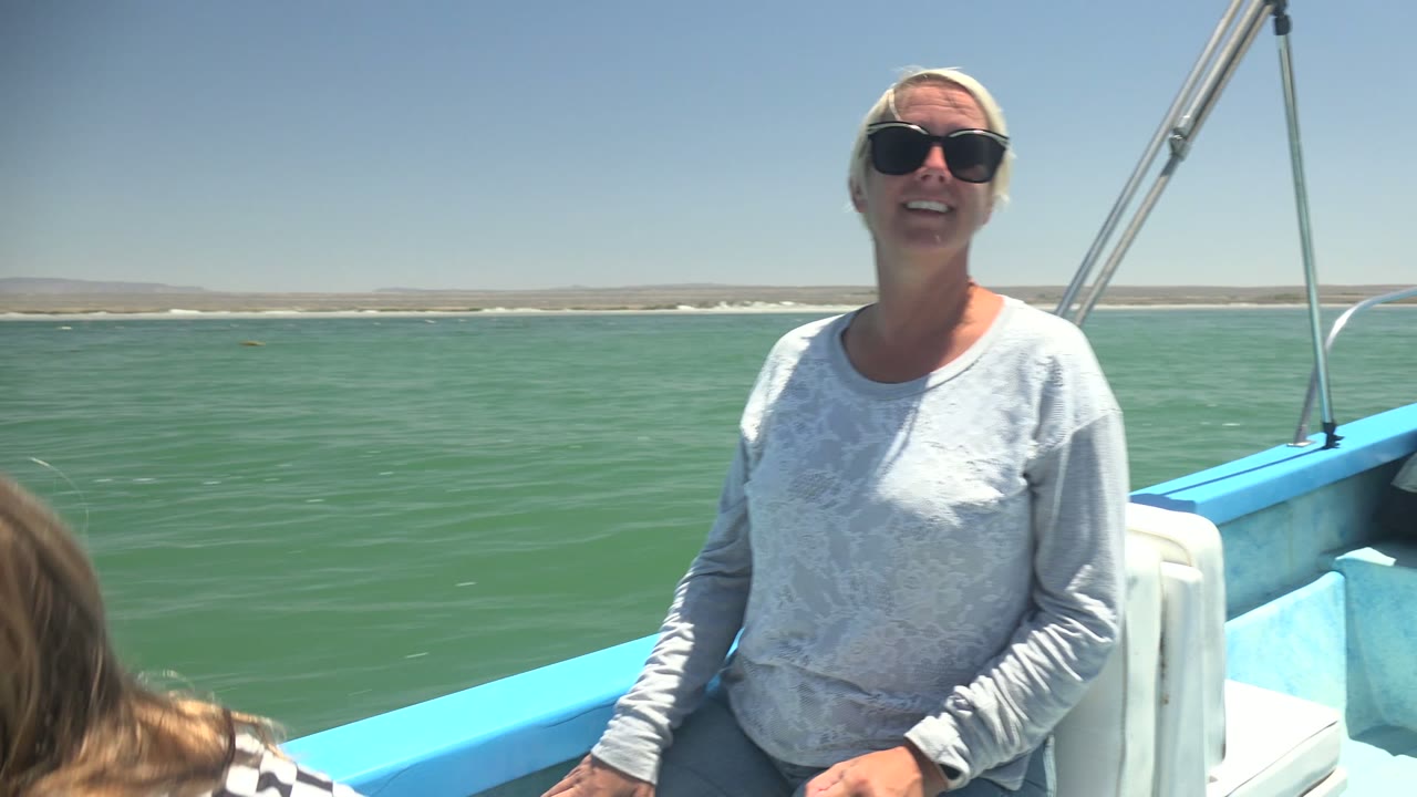 A smiling woman sits on the blue boat in La Bocana, Mexico, while the top of a child's head appears in the foreground against calm green water and a pale shore.