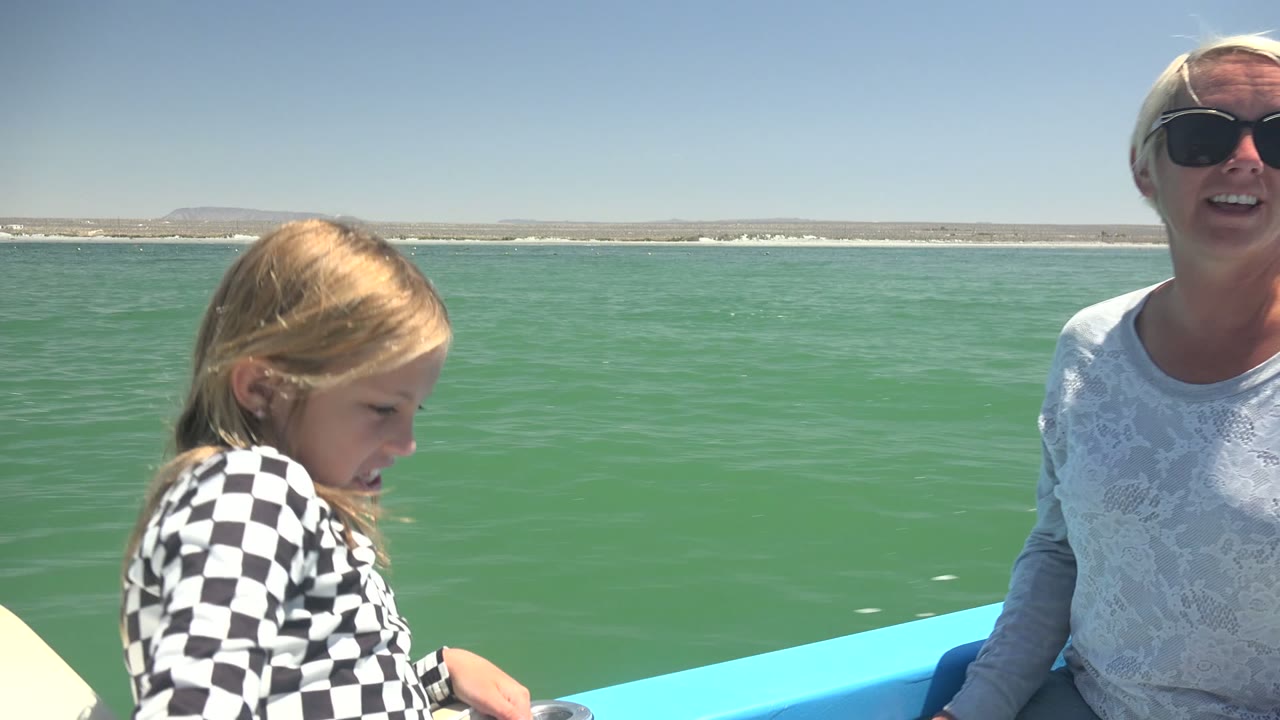 In La Bocana, Mexico, the girl smiles in profile near the boat rail while the woman in sunglasses sits partly cropped at the right edge against green-blue water and distant shore.