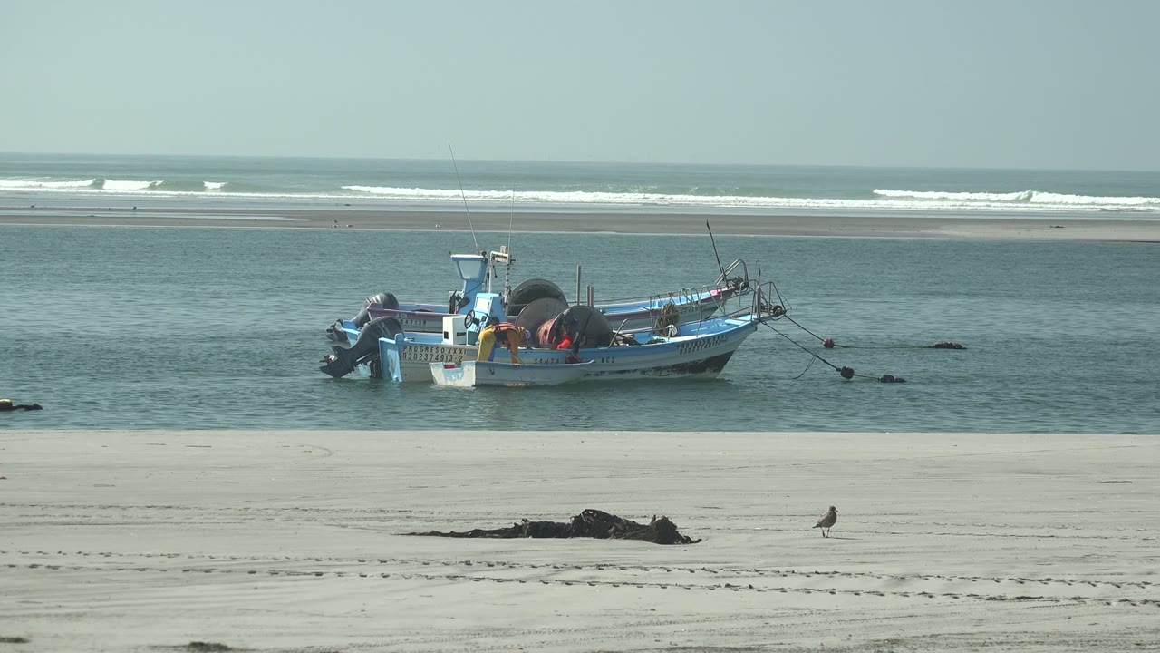 A blue fishing boat floats near the sandy shore at La Bocana, Baja California Sur, with distant white surf and no visible people.
