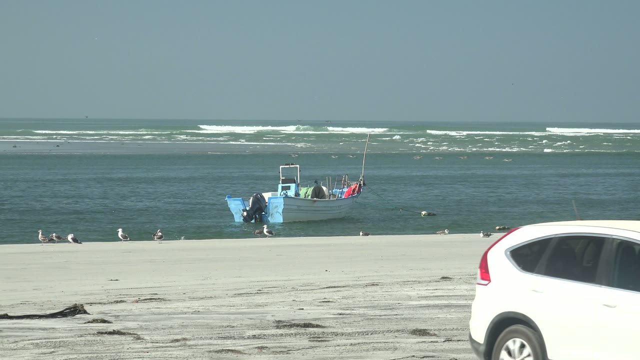 At La Bocana, a white car begins entering from the right edge while the blue boat, seabirds, pale sand, ocean, and surf remain visible behind it, with no visible people.