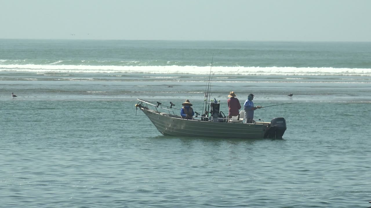 Small motorboat with people fishing in La Bocana, Baja California Sur, with rods upright, pale surf bands, and birds along the distant shore.