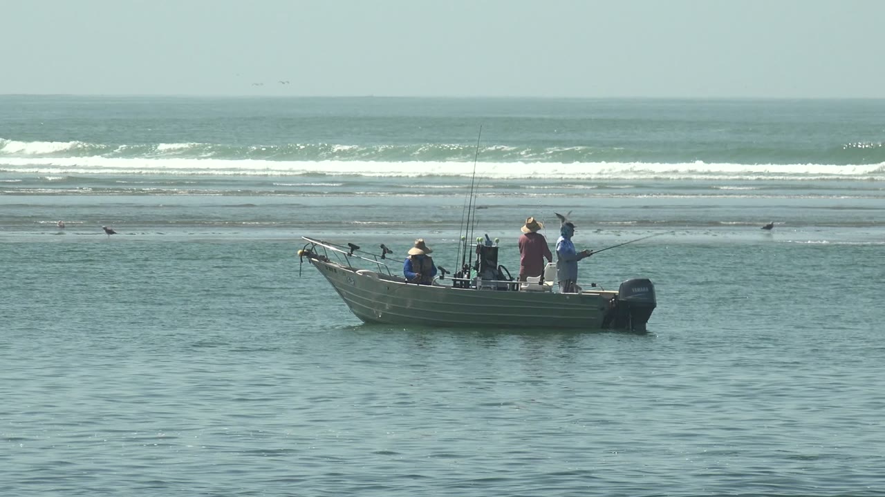 The La Bocana fishing boat remains on calm blue-gray water, with several people visible and a low line of white waves breaking across the background.