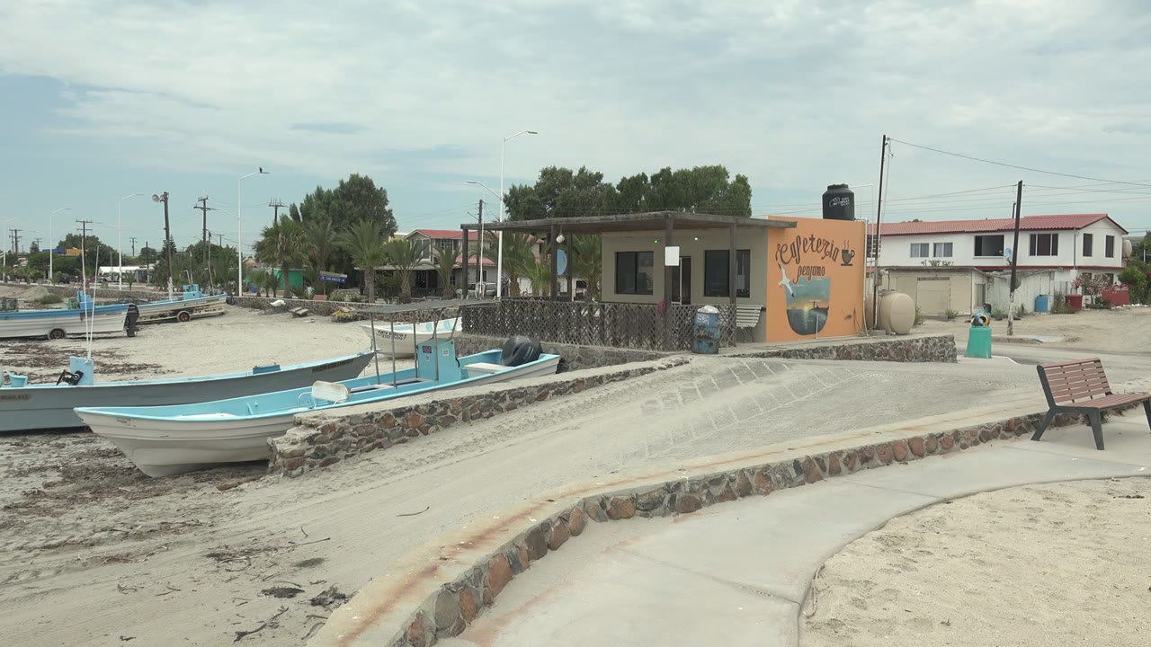 Punta Abreojos, Baja California Sur beach view with light blue and white boats on sand, a low stone wall, a bench, a cafeteria building, trees, utility poles, ocean water, cloudy sky, and no visible people.