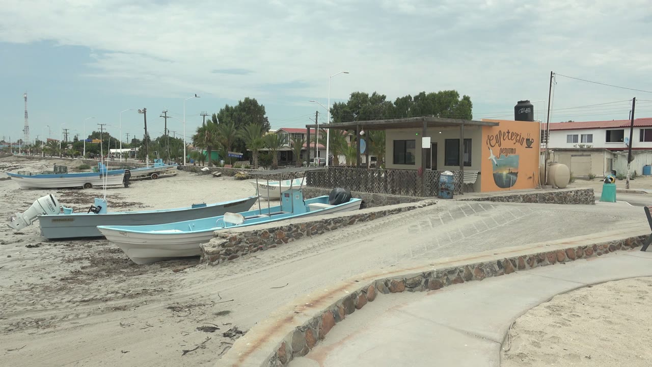 Punta Abreojos shoreline scene with boats resting on sand, a bench by a curved concrete path, a low stone wall, the cafeteria building at right, distant shoreline structures, cloudy sky, and no visible people.