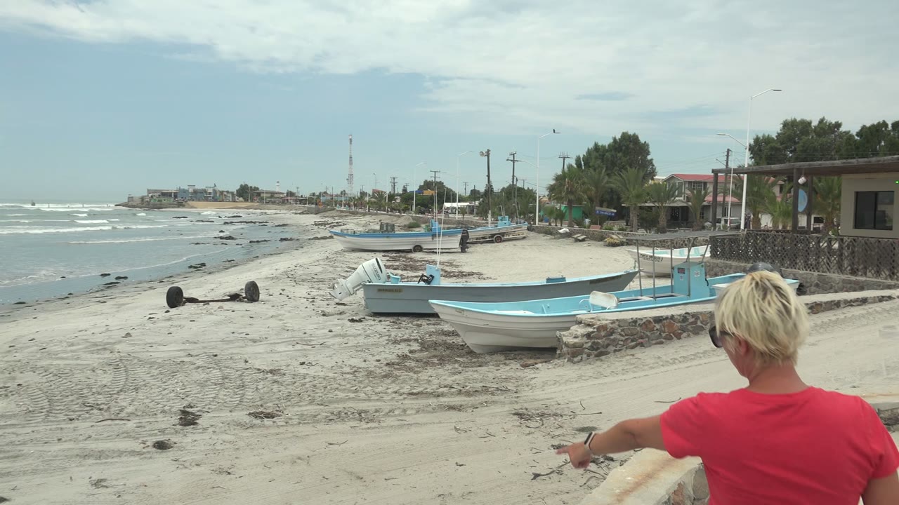 At Punta Abreojos BCS Mexico, a blonde person in a red shirt enters the foreground while blue boats rest on sand beside the ocean waves.