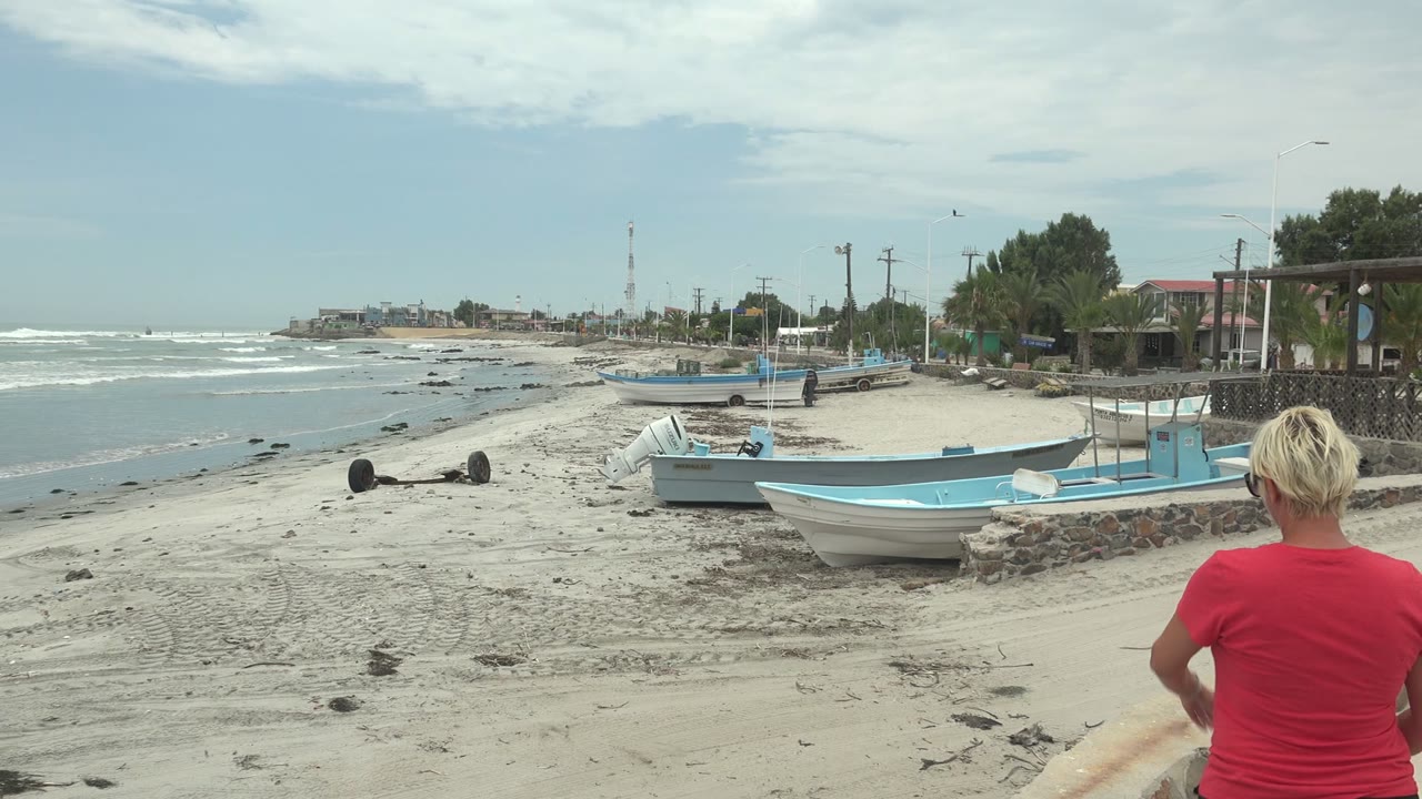 Person in a red shirt standing near the foreground with blue boats and waves behind.