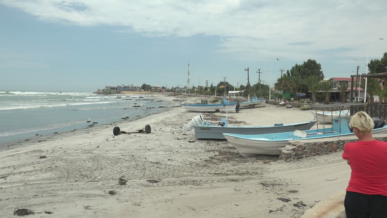 At Punta Abreojos BCS Mexico, the red-shirted beachgoer shifts to the right edge while the boats, surf, sand tracks, and distant shoreline remain visible.