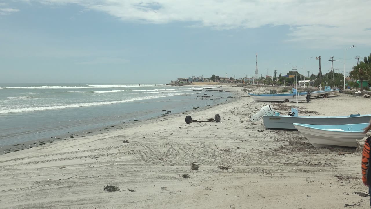 Punta Abreojos beach in Baja California Sur with pale sand, small boats, low waves, distant shoreline structures, and a person in orange partly visible near the lower right edge.