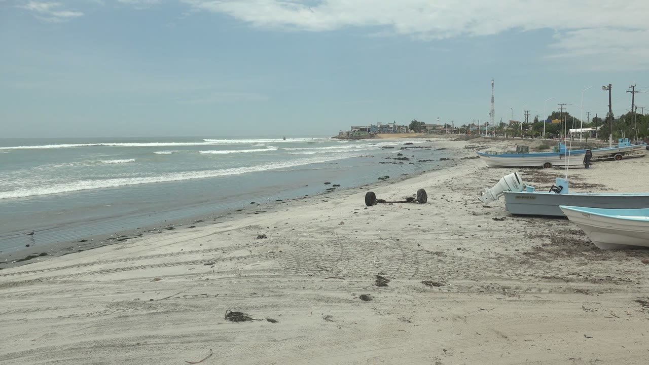 Punta Abreojos beach with no visible people, a white boat cropped at the right foreground, blue-trimmed boats farther back, and low waves breaking across the left side.