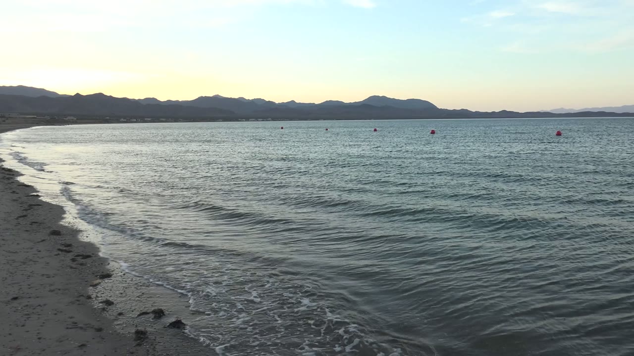 At Bahia De Los Angeles, a small wave curls toward the sandy shoreline from the left while dark mountains span the horizon under pale sky, with no visible people.