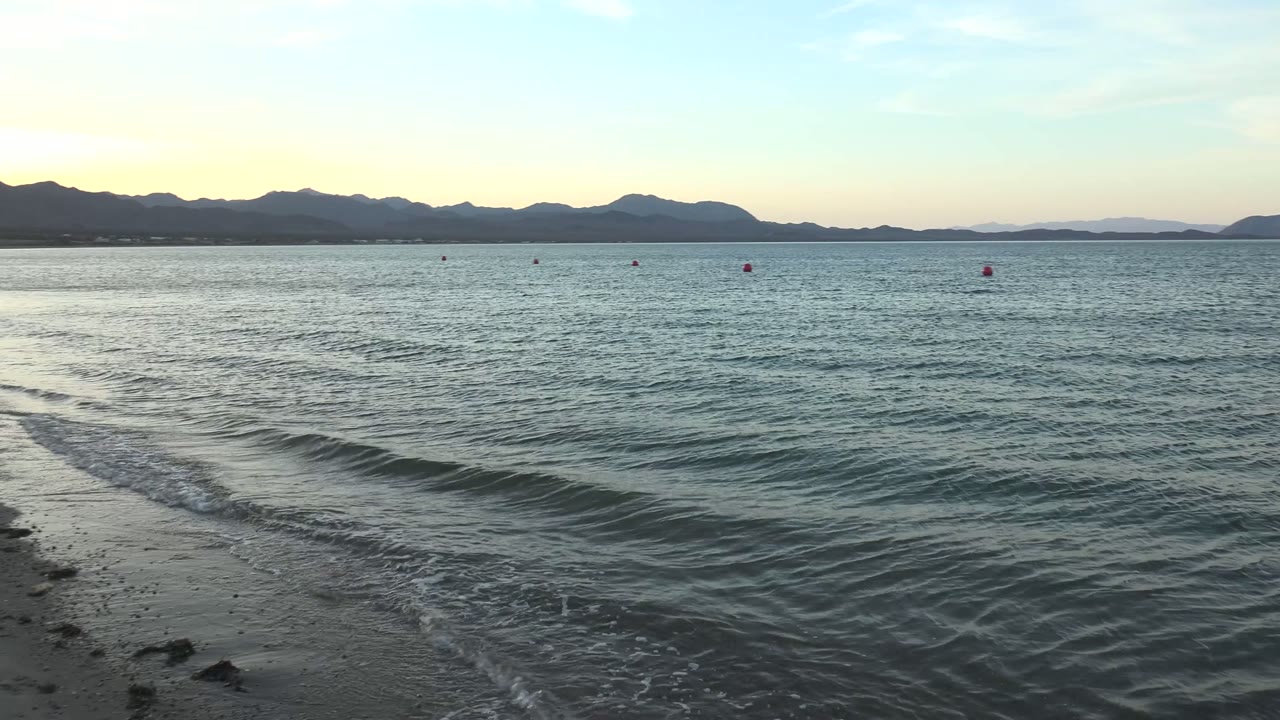 At Bahia De Los Angeles, a thin foamy wave line cuts diagonally across the foreground sand while open water, scattered buoys, and distant mountains fill the background, with no visible people.