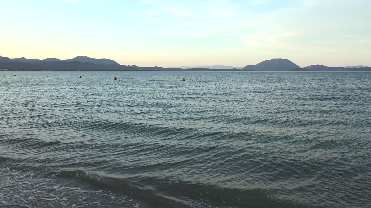 Gentle waves roll across the foreground in Bahia De Los Angeles, with small red buoys on the water, distant mountain silhouettes, pale sky, and no visible people.