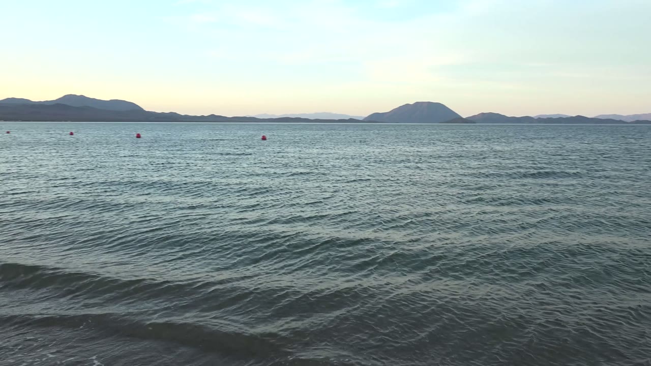 Low waves approach the sandy edge in Bahia De Los Angeles while the horizon holds dark mountains, scattered red buoys, a pale sky, and no visible people.