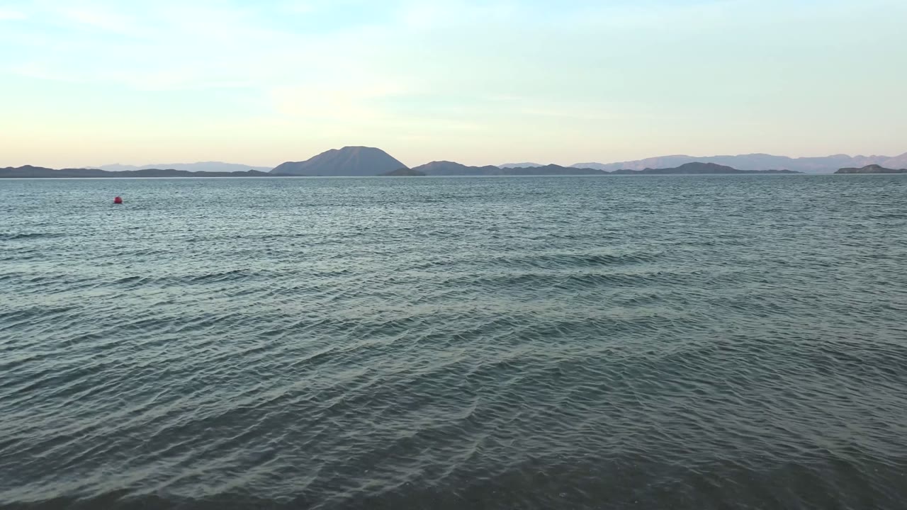 Opening view of Bahia De Los Angeles water with gentle ripples in the foreground, distant island silhouettes across the horizon, small red buoys, pale sky, and no visible people.
