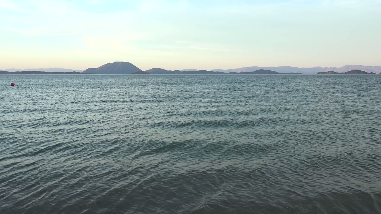 Mid-clip view of Bahia De Los Angeles with rolling ripples in the foreground, open sea in the middle distance, dark island shapes on the horizon, soft sky color, and no visible people.