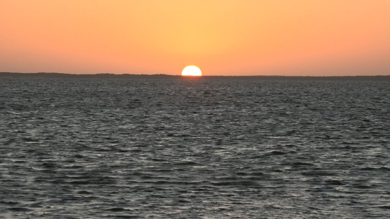 The sun rests just above the distant horizon over dark rippled water in San Carlos, Baja California Sur, with an orange sky and no visible people.