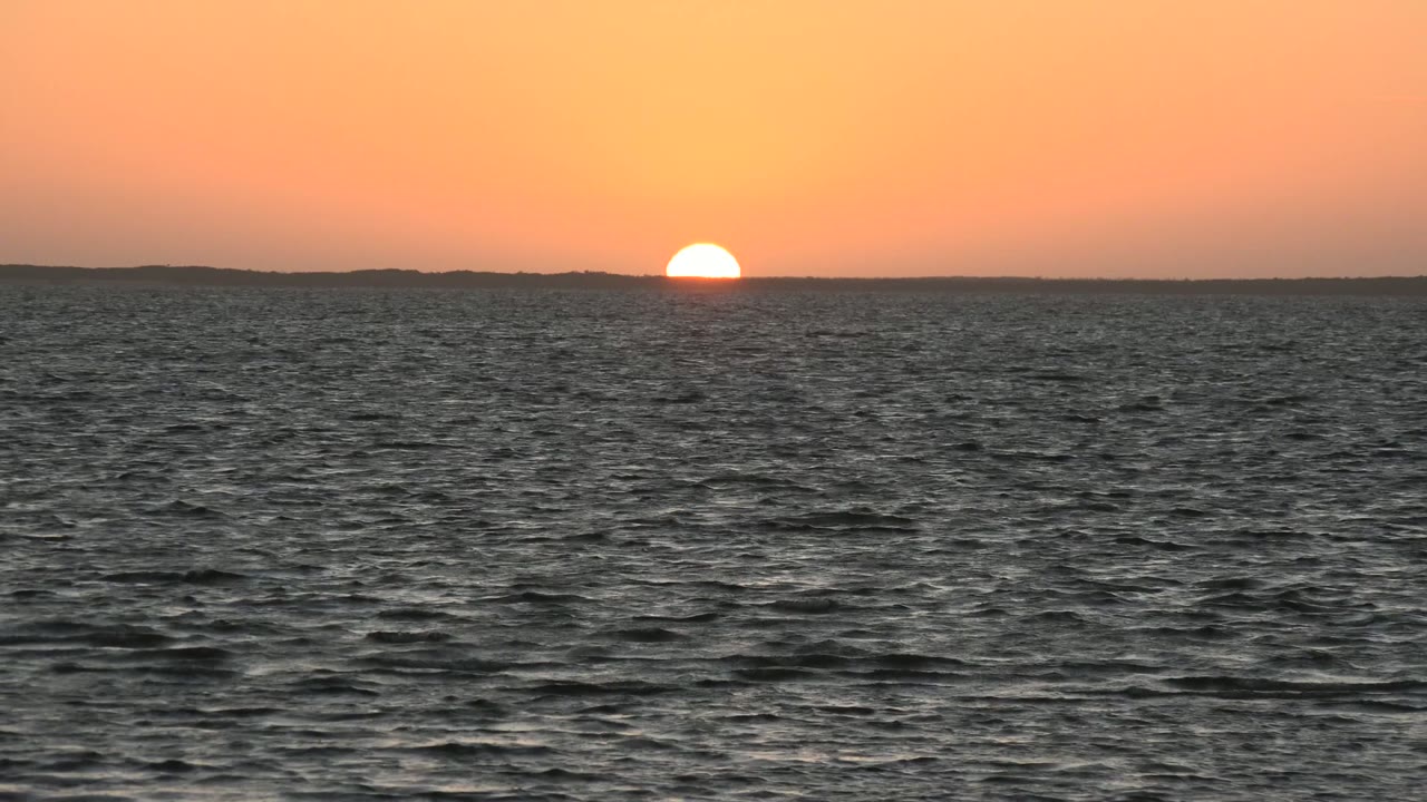 At 0:00 in San Carlos, Baja California Sur, the sun sits half above a distant dark horizon with rippled ocean water in front and no visible people.
