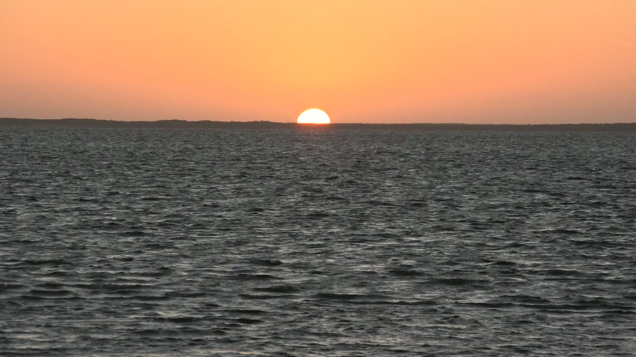 At 0:02, the orange sky glows above the distant land line while the foreground sea shows uneven ripples and no visible people.