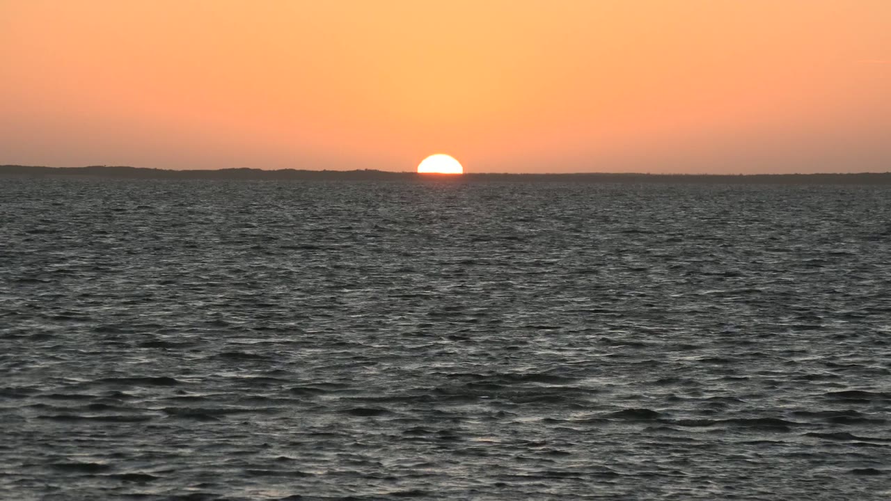 At 0:00, the sun is half visible above the distant land horizon in San Carlos, Baja California Sur, with dark rippled ocean water below and no visible people.