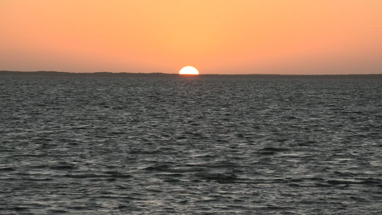 At 0:02, the sun remains a bright half circle against the orange sky over the San Carlos horizon, with rippled ocean in front and no visible people.