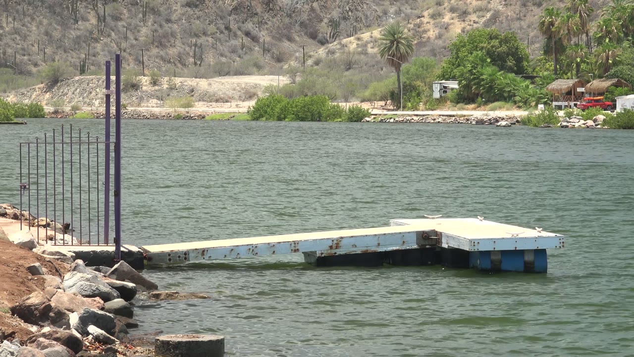 A pale floating dock extends into green water in Mulege, Baja California Sur, with rocks at left, vertical posts, far trees, a white trailer, hills, and no visible people.
