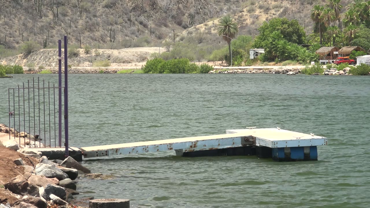 The dock remains centered over the rippling Mulege water, with the red vehicle, white trailer, trees, palms, and dry hillside visible across the far bank, and no visible people.