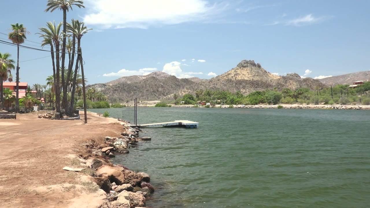 Mulege waterfront frame showing more open water to the right, rocks along the near bank, tall palms and a small red-roofed building at left, dry mountains beyond, and no visible people.