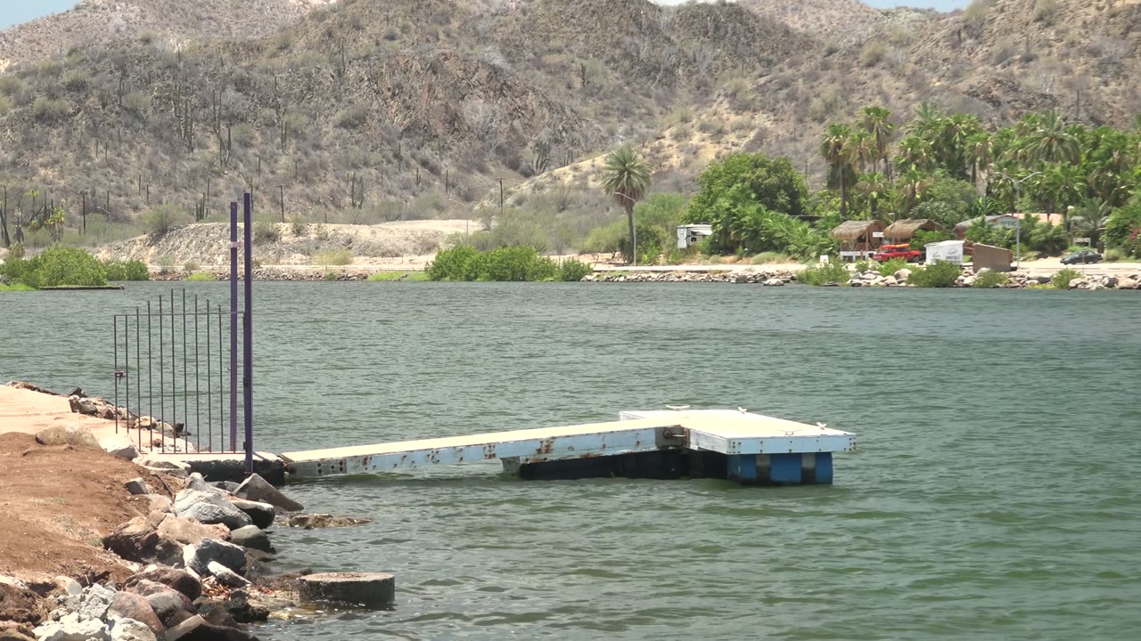 A floating dock extends from a rocky shoreline into green water in Mulege, Baja California Sur, with dry mountains and palms across the background and no visible people.