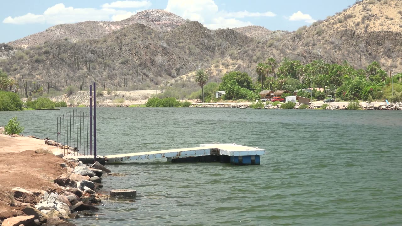 A low dock projects from the rocky bank into the water, with the Mulege mountain ridge, palms, and small shore structures visible under scattered clouds, no visible people.