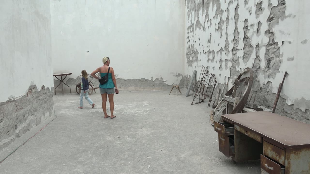 At 0:00 in Mulege, Mexico, a child stands near the left wall while a woman faces away in a worn concrete courtyard with peeling white paint and old furniture along the right side.