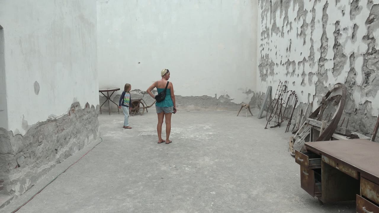 At 0:02 in Mulege, Mexico, the child and woman are separated across the courtyard, with the wheelbarrow near the rear wall and stacked furniture and tools visible on the right.