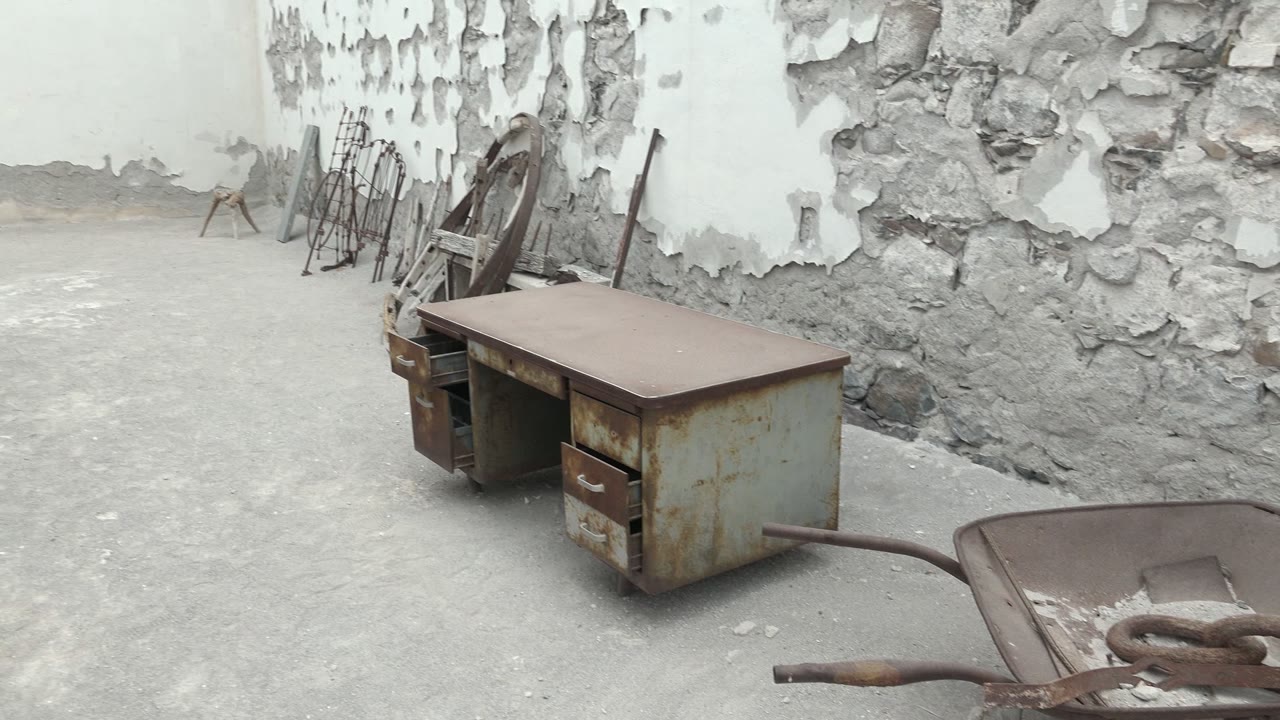 A Mulege courtyard frame shows a rusty metal desk, a wheelbarrow handle in the foreground, exposed stone under peeling plaster, stacked chairs along the wall, and no visible people.
