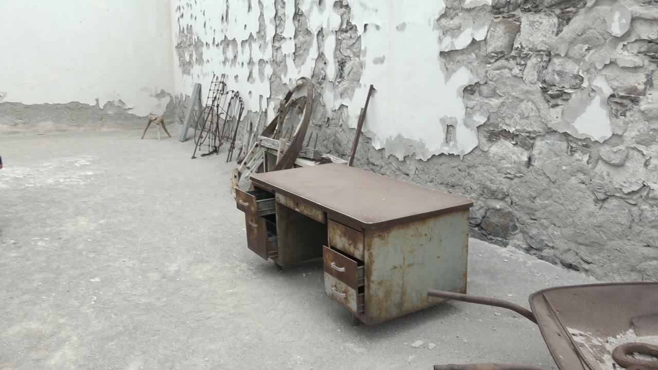Rusty desk, wheelbarrow, chairs, and peeling wall in an empty courtyard.
