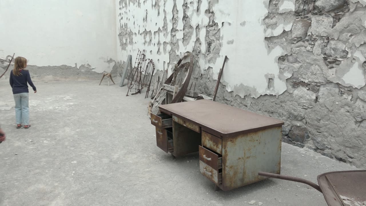 A girl walks away through the Mulege courtyard near a peeling wall, with a rusty desk, wheelbarrow edge, stacked chairs, and open concrete space around her.
