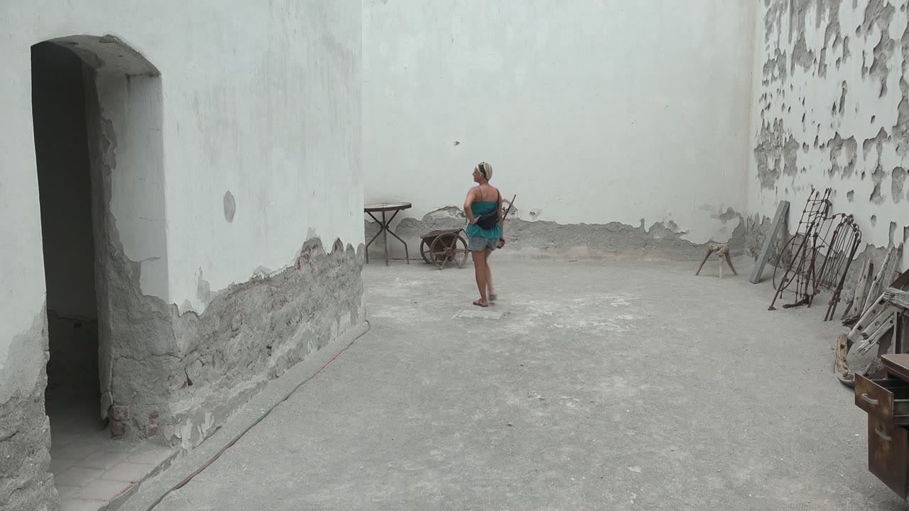 The woman appears farther from the camera in the Mulege courtyard, centered against a pale wall with a wheelbarrow and small table to her left and stacked items to her right.