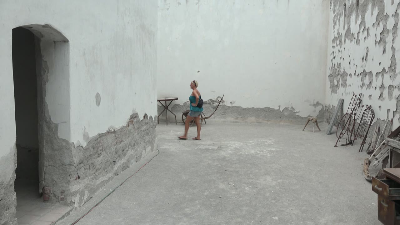 Woman walking left across the courtyard beside peeling white walls.