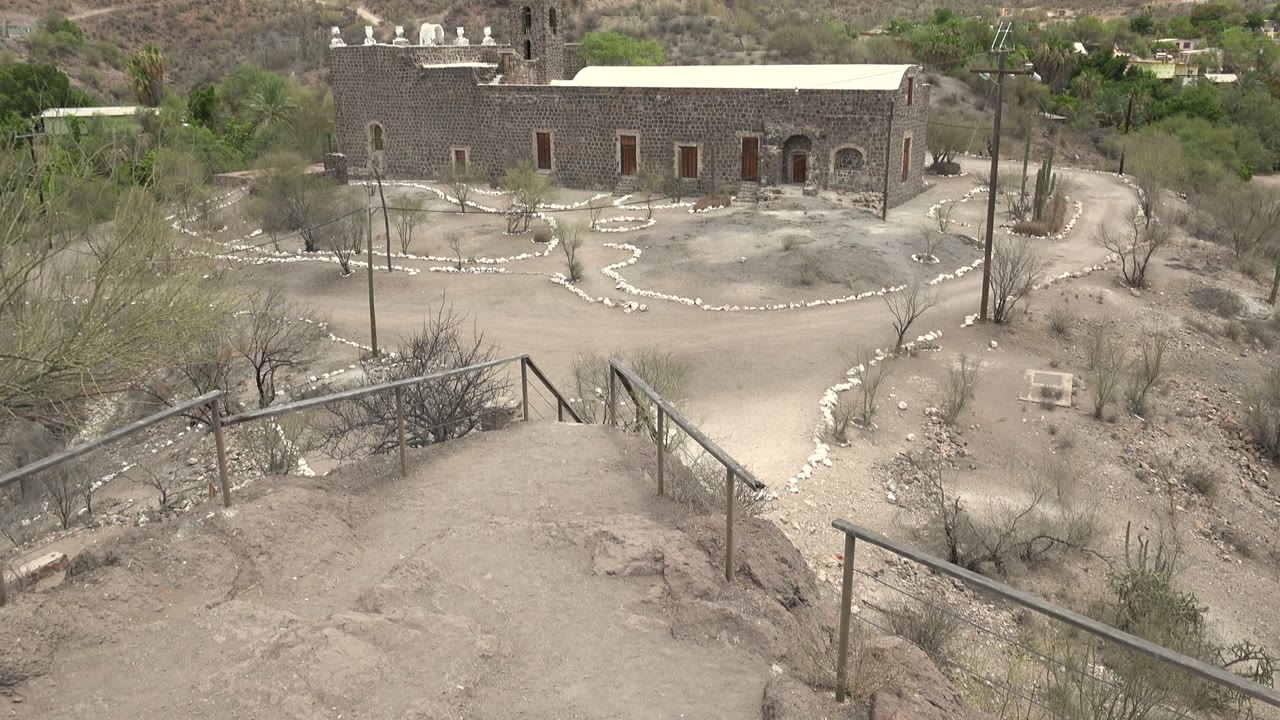 Wide view of a stone mission building in Mulege, Baja California Sur, with a sandy courtyard, white stone path edging, railings in the foreground, hills behind, and no visible people.