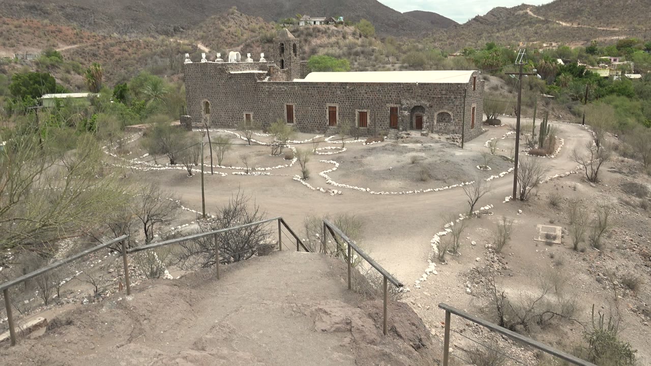A slightly shifted wide frame shows the stone mission building in Mulege with more dry brush at the right edge, utility poles beside the courtyard, low hills in back, and no visible people.