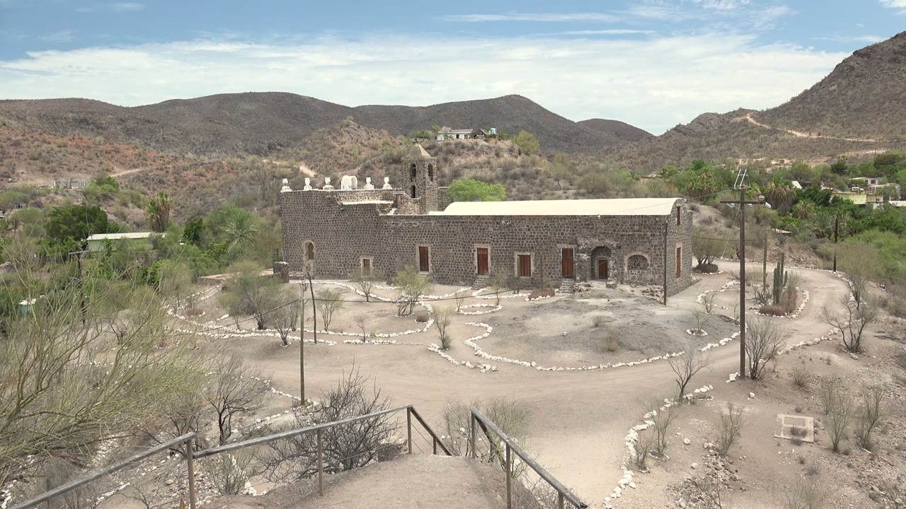 Wide view of a stone mission building in Mulege, Baja California Sur, with sandy ground, low rock borders, foreground brush, distant hills, and no visible people.