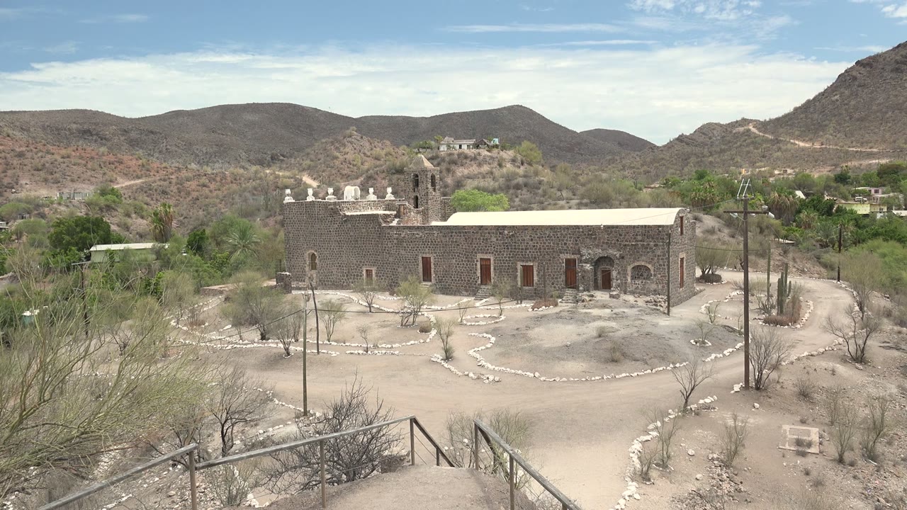 Mission building and cacti below a blue sky with mountain ridges in back.